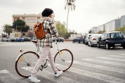 An interracial hipster is pushing his bicycle across the zebra while visiting Spain.