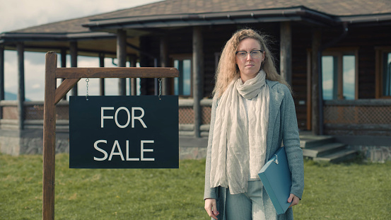 Female real estate agent stand near for sale sign and looks at camera. real estate agent waits for visitors. Stylish house with traditional architecture in mountains. Residential property on sale concept.