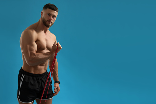 Muscular man exercising with elastic resistance band on light blue background. Space for text
