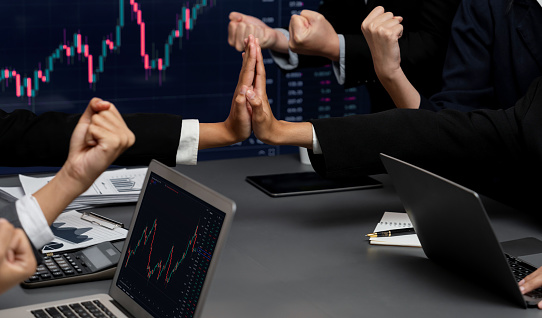 Group of stock trading investors in brokerage firm company celebrating together after achieving profitable gain from selling stocks, expressing joy through celebratory high five gesture. Trailblazing