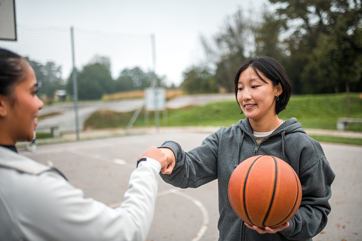 A Hispanic female and Asian female share a fist pump after their basketball game, highlighting the diverse bond of their friendship.