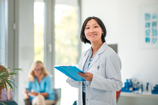 A mid adult Asian female doctor, wearing a lab coat, standing in a waiting room, holding a clipboard.