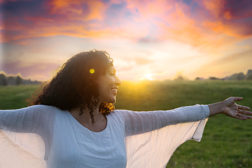 Happy smiling mid adult latin woman spreading hands and running in summer field at sunset, medium sjot