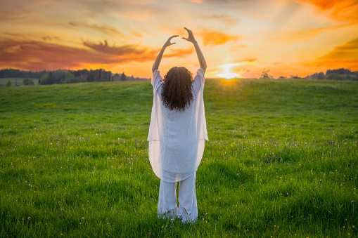Latin woman raising hands and standing in summer field at sunset, wide shot back view