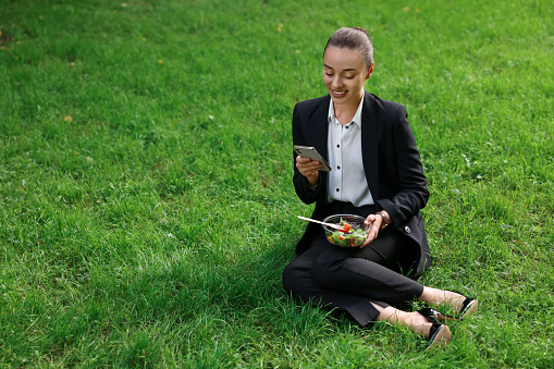 Lunch time. Happy businesswoman with container of salad using smartphone on green grass outdoors, space for text