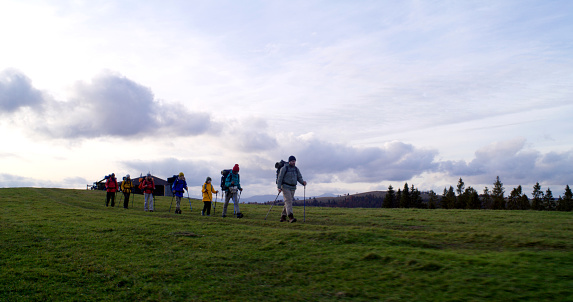 Full shot of diverse group of tourists walking on trail after sleepover in big house. Hiking buddies during trip or trek to mountains on their vacation in autumn. Tourism and active leisure concept.