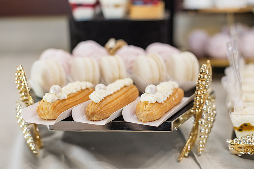 Delicate eclairs topped with cream and pearls, alongside soft meringues, presented on sophisticated dessert tray with golden accents
