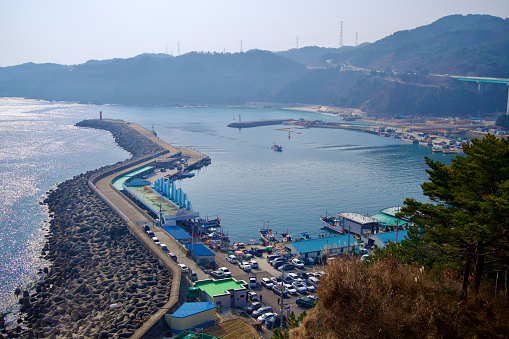 Samcheok City, South Korea - December 28, 2023: Elevated view of Imwon Port from a seaside mountain, capturing a boat sailing into port, the long breakwater, and the market area below.