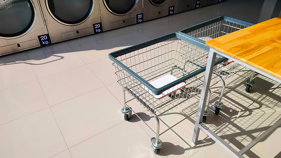 Two laundry basket trolley and table on tile floor with row of vending washing machines and clothes dryer inside of modern laundromat shop, high angle view with copy space