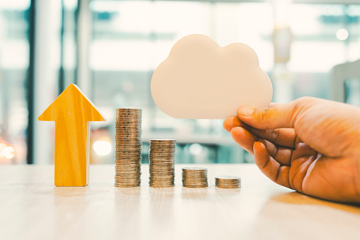 A pile of money arranged from low to high with an arrow pointing upwards, together with a child's hand holding a wooden cloud model, It is a concept of cryptocurrency that is deposited in the Cloud.