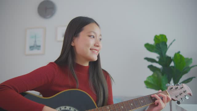Asian woman using laptop learn to play guitar in living room at home. Attractive beautiful young girl student feel happy and relax enjoy studying how to use instrument on table during weekend in house