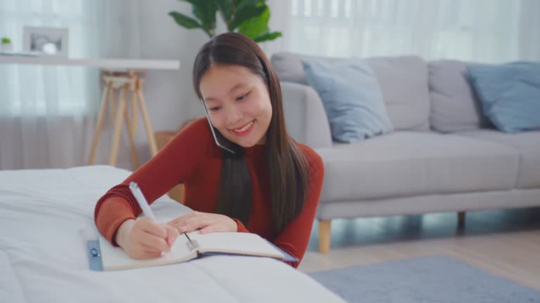 Asian young beautiful woman taking note on book while talking on phone. Attractive girl using smartphone call and writing note on study record ​document paper on working table in living room at home.