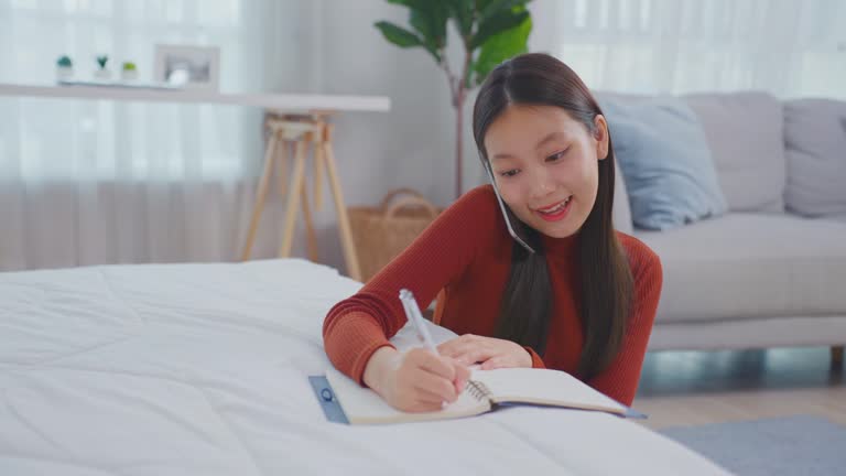 Asian young beautiful woman taking note on book while talking on phone. Attractive girl using smartphone call and writing note on study record ​document paper on working table in living room at home.