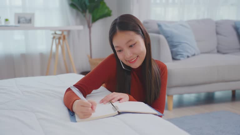 Asian young beautiful woman taking note on book while talking on phone. Attractive girl using smartphone call and writing note on study record ​document paper on working table in living room at home.