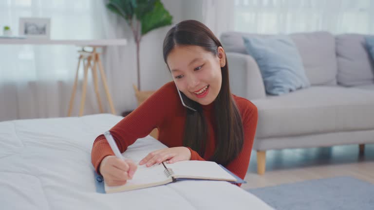 Asian young beautiful woman taking note on book while talking on phone. Attractive girl using smartphone call and writing note on study record ​document paper on working table in living room at home.