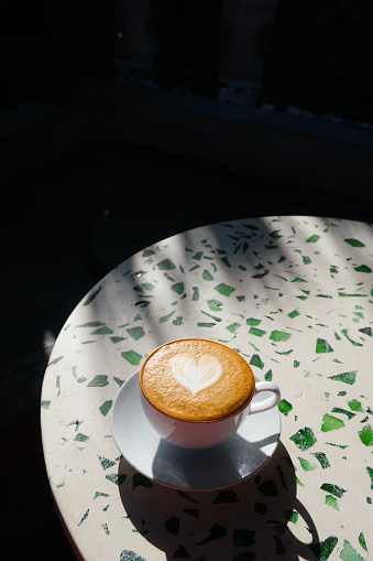 A white cup of latte casting a heart shadow on a terrazzo table in sunlight