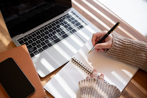 A top-view image of a woman in a cosy knitted sweater is holding a pen, writing something, or taking notes in her notebook while working on her laptop indoors.