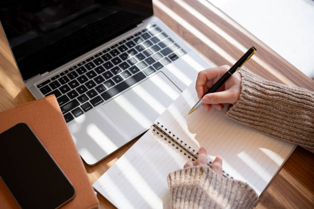 A top-view image of a woman is taking notes in her notebook while working on her laptop indoors. A top-view image of a woman in a cosy knitted sweater is holding a pen, writing something, or taking notes in her notebook while working on her laptop indoors. books student stock pictures, royalty-free photos & images
