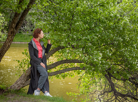 A mature woman sat down on a branch of a flowering tree bent over the water and inhales the fragrance of its flowers. Springtime in the park