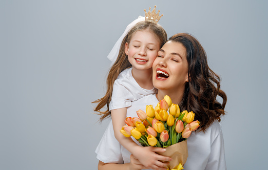Happy women's day. Child is congratulating mom and giving her yellow flowers. Mum and girl smiling on light grey background. Family holiday and togetherness.
