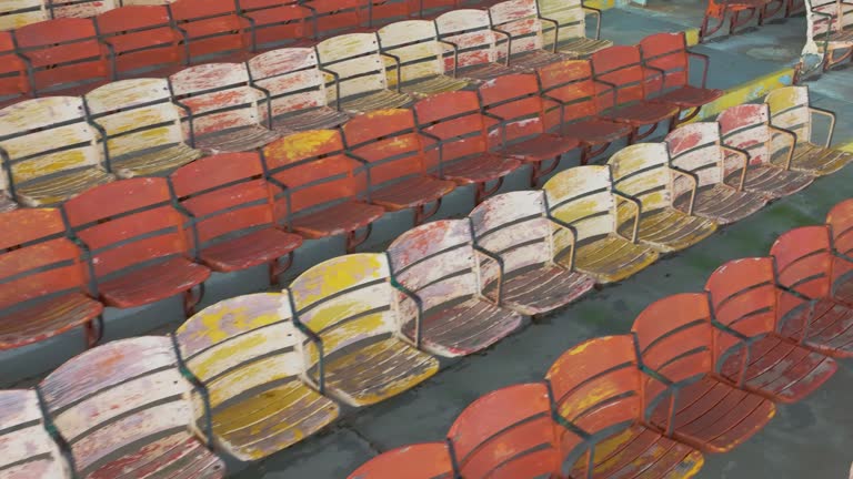 Drone, aerial, video of old faded orange and yellow painted wooden seats at a abandoned a stadium grandstand.