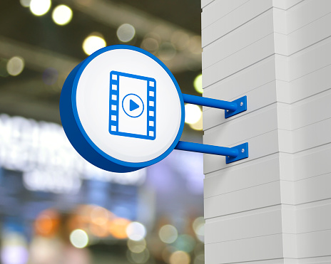 Play button with movie icon on hanging blue rounded signboard over blur light and shadow of shopping mall, Business cinema online concept, 3D rendering