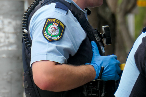 Officers of the New South Wales Police in attendance at an incident on Bondi Road, Bondi where one man was handcuffed. They are wearing light-armour vests.  On the left is a streetlight post with remains of sticky tape from advertising posters. This image was taken on a hot, humid and sunny morning on 26 January 2024.