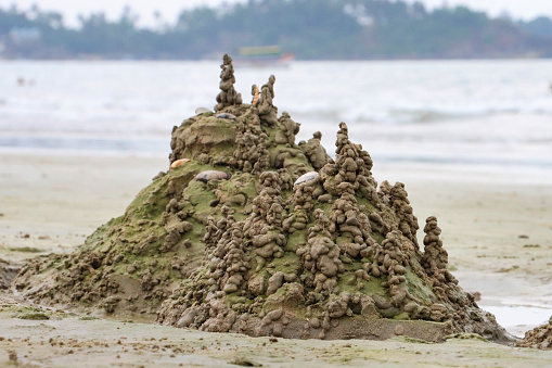 Stock photo showing close-up view of a sandcastle surrounded by water filled moat built on a sunny, golden sandy beach with sea at low tide in the background. This form of sand castle is made by dribbling fistfuls of wet sand to form towers.