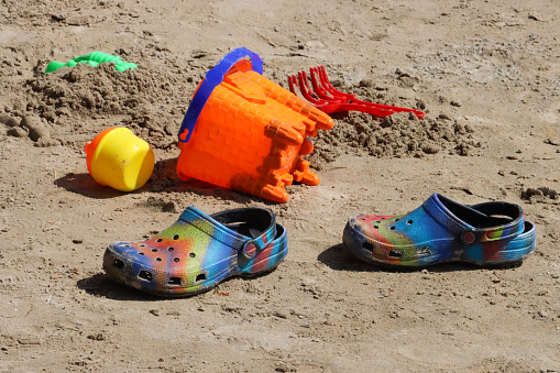 Stock photo showing a collection of multicoloured, plastic beach toys on sandy beach near water's edge of sea. Toys include a castle shaped bucket, rake, sand moulds and a watering can.