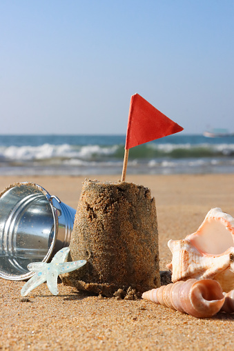 Stock photo showing close-up view of a metal bucket, pile of seashells and starfish besides a sandcastle decorated with a red triangular flag, on a sunny, golden sandy beach with sea at low tide in the background. Summer holiday, tourism and activities concept.