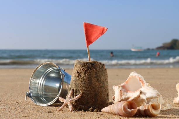 istock Close-up image of sandcastle decorated with triangular, red flag, metal bucket on sunny sandy beach, pile of seashells, waves breaking at low tide, sea and coastline, focus on foreground, Summer holiday and tourism concept, copy space 1961824763