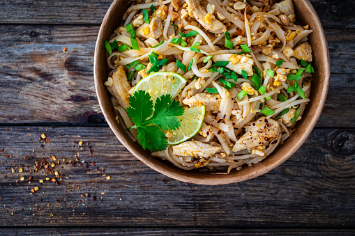 Pad Thai with chicken nuggets and rice noodles in peanut and tamarind sauce on wooden table in eco bowl to go