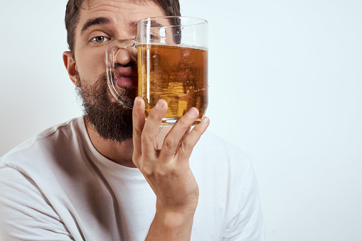 A bearded man with a mug of beer on a light background in a white T-shirt cropped view of an alcoholic drink. High quality photo