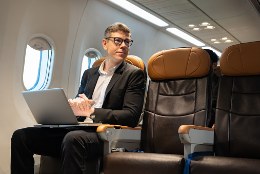 Young attractive businessman with eyeglasses working with laptop computer while sitting in airplane cabin. Internet connection on board service and business travel concept.