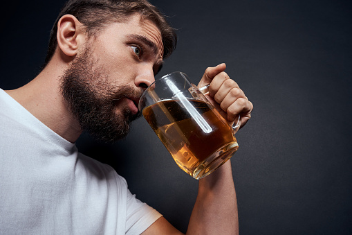 man with a mug of beer in a white t-shirt emotions lifestyle drunk on a dark isolated background. High quality photo