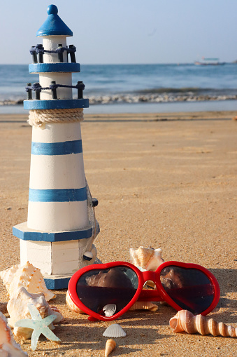 Stock photo showing close-up view of blue and white striped model lighthouse and red, heart-shaped sunglasses on a sunny, golden sandy beach besides a pile of seashells, with sea at low tide in the background.