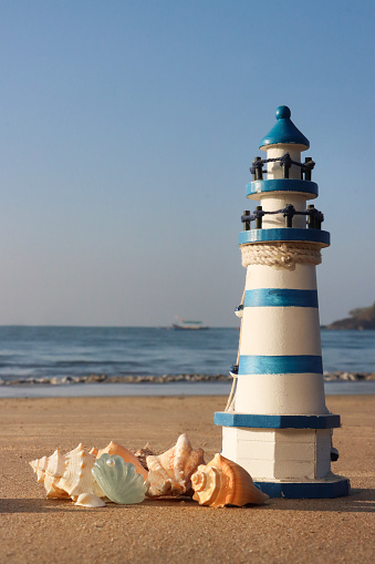 Stock photo showing close-up view of blue and white striped model lighthouse on a sunny, golden sandy beach besides a pile of seashells, with sea at low tide in the background.