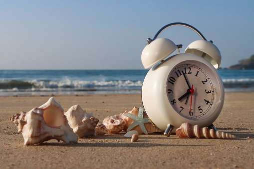 Stock photo showing close-up view of retro, double bell, analogue alarm clock, with black numerals, hour and minute hands, on a sunny, golden sandy beach besides a pile of seashells and starfish, with sea at low tide in the background.  Summer daylight savings concept