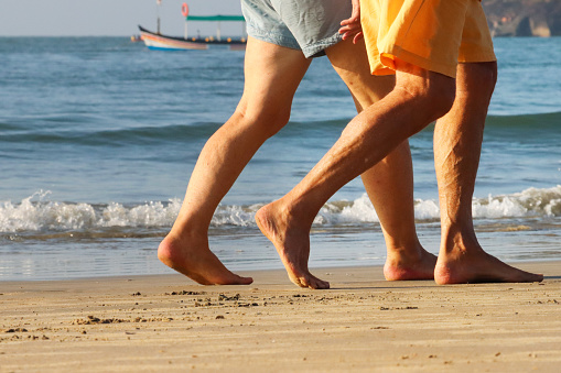 Stock photo of unrecognisable people walking barefoot along sunny, golden sandy beach with sea at low tide in the background.