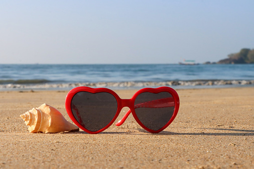Stock photo showing close-up view of red, heart-shaped sunglasses besides a helix seashell on a sunny, golden sandy beach with sea at low tide in the background.
