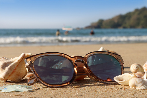 Stock photo showing close-up view of tinted, mirrored sunglasses besides a pile of seashells on a sunny, golden sandy beach with sea at low tide in the background.