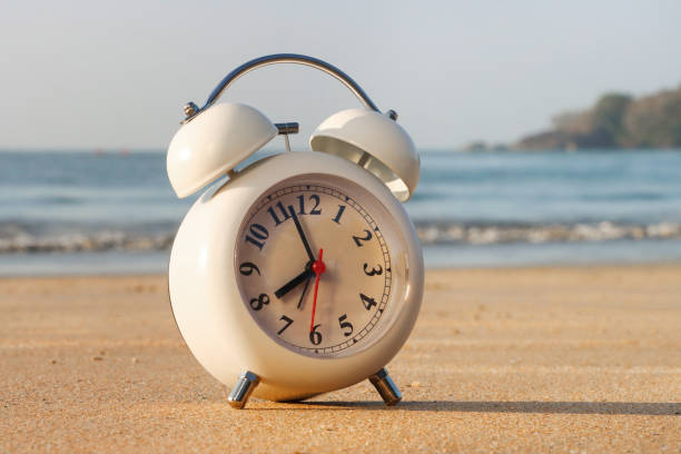 Stock photo showing close-up view of retro, double bell, analogue alarm clock, with black numerals, hour and minute hands, on a sunny, golden sandy beach with sea at low tide in the background.  Summer daylight savings concept