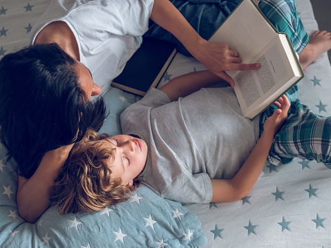 Top view of preteen boy and mom reading interesting book while lying on bed