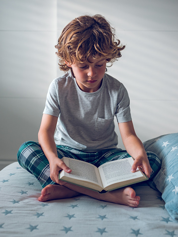 Full body of focused preteen boy in sleepwear sitting with crossed legs on soft bed and reading exciting story in book