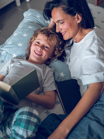From above of happy preteen boy lying on bed with mother and reading book