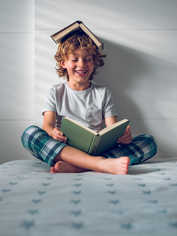 Full body of smiling little boy in nightwear sitting on bed with crossed legs and reading interesting story with book covering head