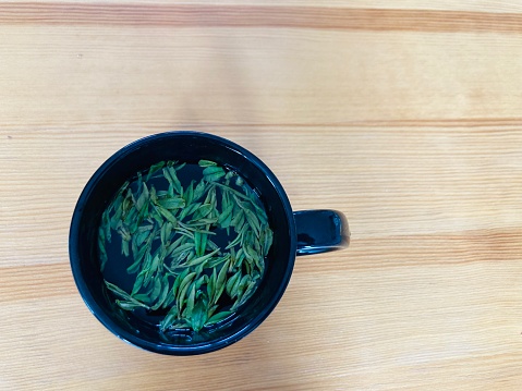 hot natural dried green tea leaves in a black cup on a light brown wooden table with the copy space on the right side of the frame