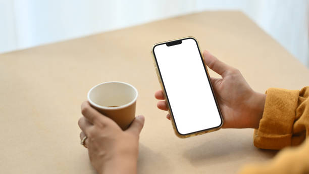 istock Closeup woman using mobile phone and drinking coffee at desk. Empty screen for advertising or message 1961912086