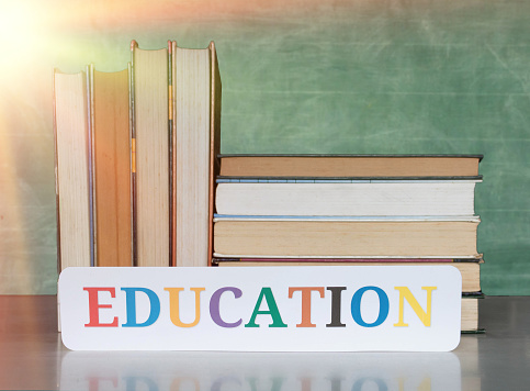 stack of textbooks and education card on desk over blurred blackboard in classroom