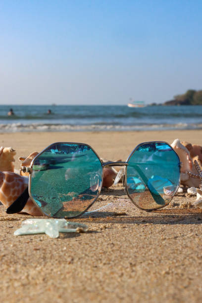 Stock photo showing close-up view of tinted, mirrored sunglasses besides a pile of seashells on a sunny, golden sandy beach with sea at low tide in the background.
